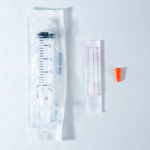 A packaged syringe, a packaged needle, and an orange needle cap on a white surface.