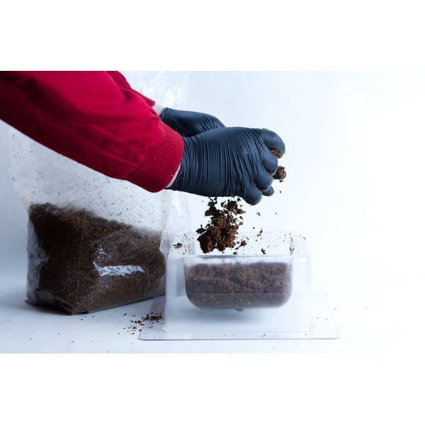 A person in black gloves adds soil to a clear container, with a soil bag nearby—showing how to use the Mycelium Mould Growth Form - Plant Pot Heavy Duty (Reusable) for optimal results.