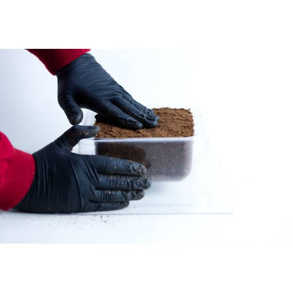 Hands in black gloves pressing soil into a clear plastic container on a white surface, preparing it for the Mycelium Mould Growth Form – Plant Pot Heavy Duty (Reusable).