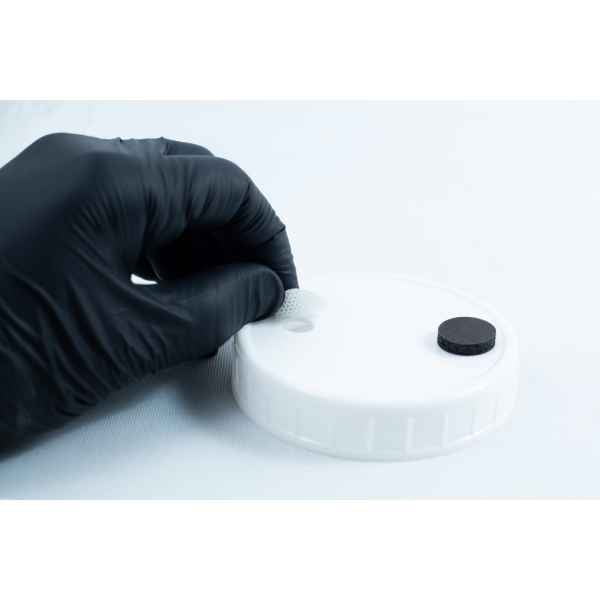 A gloved hand positions a mesh disc into a hole on a white lid beside a black disc, preparing the area for adhesion using Primer 94 multisurface for 3M glue 10ml.
