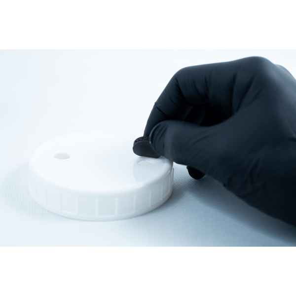 A gloved hand applies Primer 94 multisurface for 3M glue 10ML onto a round white plastic lid to prepare it for optimal tape adhesion.
