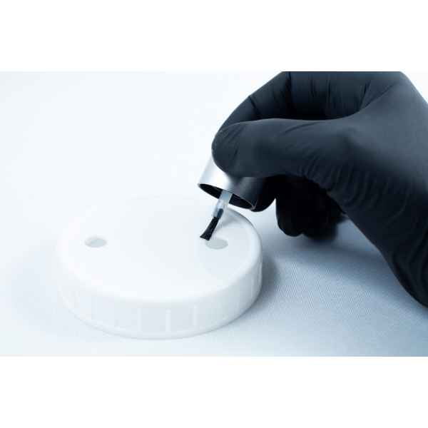 A gloved hand uses a brush to apply Primer 94 multisurface for 3M glue 10ml onto a white plastic lid with two holes, preparing it for adhesive application.