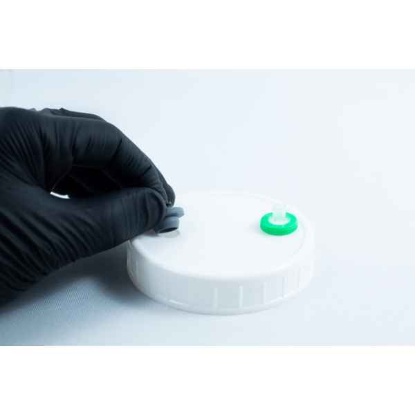 A gloved hand holds the grey cap of Primer 94 multisurface for 3M glue 10ML near a white plastic lid with a green port, commonly used as a tape primer on clean white surfaces.