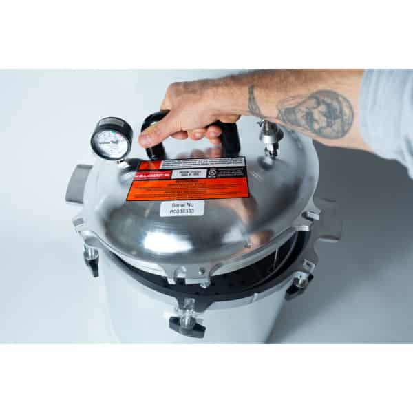 A tattooed hand holding the lid of a stainless steel pressure cooker with a gauge and warning labels.