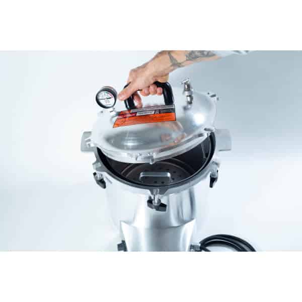 A hand lifts the lid of a stainless steel pressure cooker with a gauge and safety label on top.