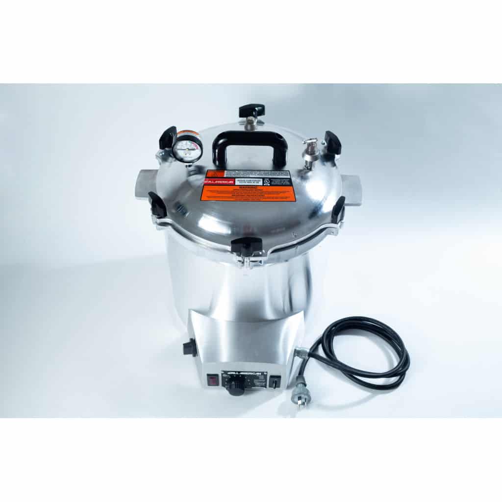 A stainless steel pressure cooker with a gauge, black handles, and an attached power lead on a white background.