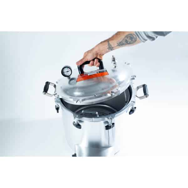 A hand lifts the lid of a stainless steel pressure cooker with a gauge on top.