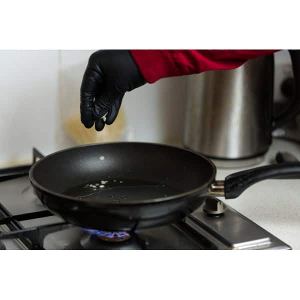 A gloved hand sprinkles ingredients from the Mushroom Log Kit | Mushroom Inoculation Kit (Sawdust Log) into a frying pan with oil on a lit gas hob.