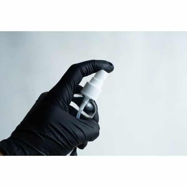 A hand in a black glove holds a 100ml spray bottle of 70% isopropyl alcohol against a plain, light background.