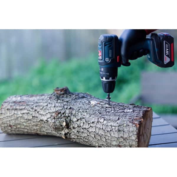 A gloved hand uses a power drill to drive a screw into a log on an outdoor table.