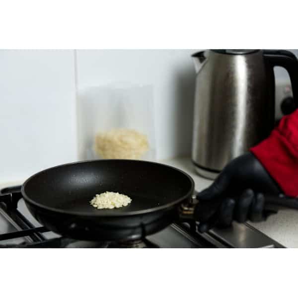 A hand in a black glove holds a pan with rice cooking on a hob next to a metal electric kettle.