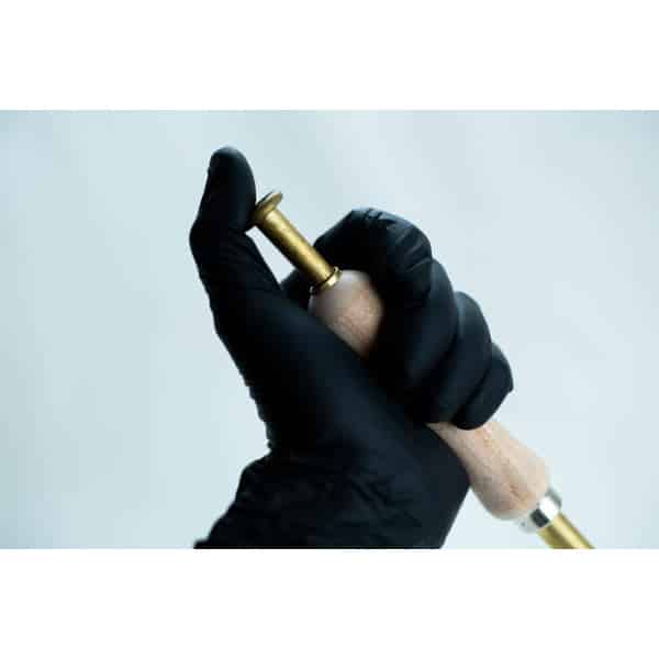 A hand in a black glove holding a brass and wooden tool against a plain light background.