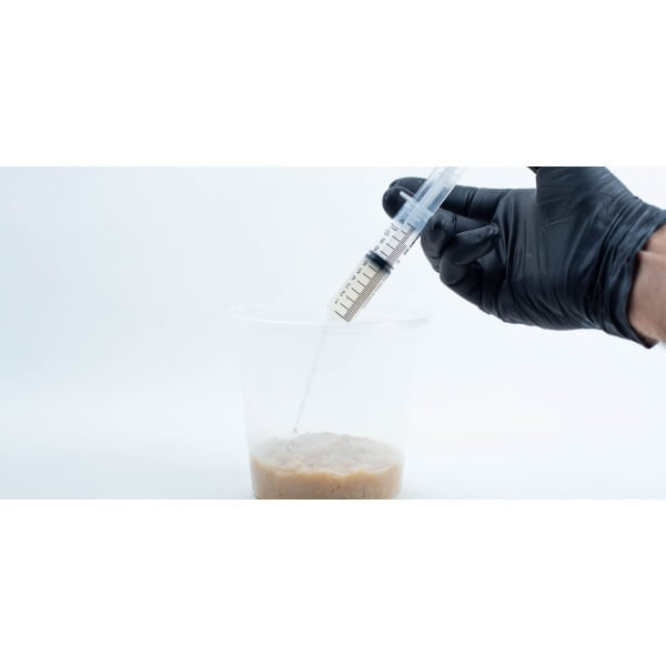 A gloved hand holds a syringe above a plastic container with a beige, lumpy liquid inside.