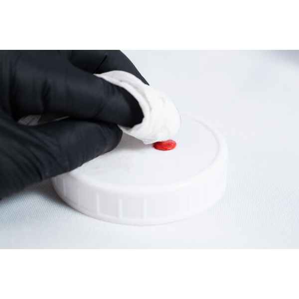 A gloved hand wipes a red stain from a white plastic lid with a tissue, ensuring the surface is clean before applying High Temperature RTV Silicone-Red.