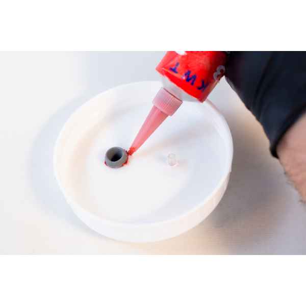 A person dispenses High Temperature RTV Silicone-Red through a nozzle into a small hole of a white circular container, preparing it for sealing.