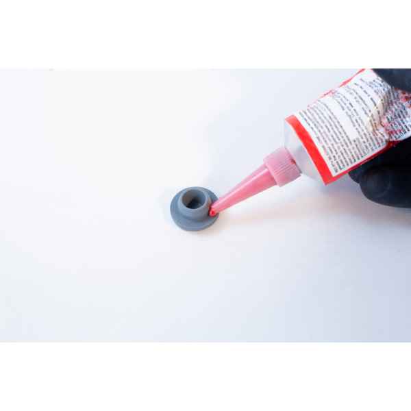 A hand applies High Temperature RTV Silicone-Red from a tube onto a small grey plastic part placed on a white surface.