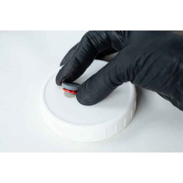 A gloved hand holds a small round object above a white plastic lid, ready to apply High Temperature RTV Silicone-Red on a light surface.