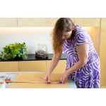 Woman in a purple dress measures cardboard with a ruler and pencil on a table in a modern kitchen.