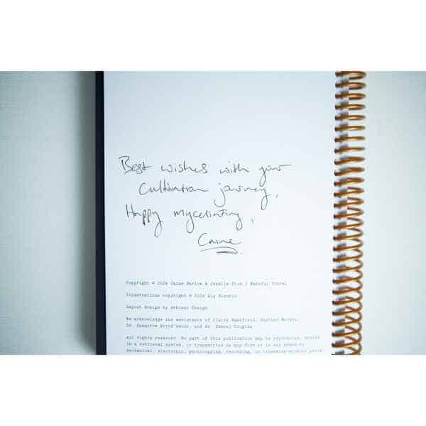 A handwritten note on a book page reads: Best wishes with your cultivation journey, happy myceliating, Carrie—inside the "Wakeful Mushroom Cultivation Journal" by Australian mycologist Caine Barlow.