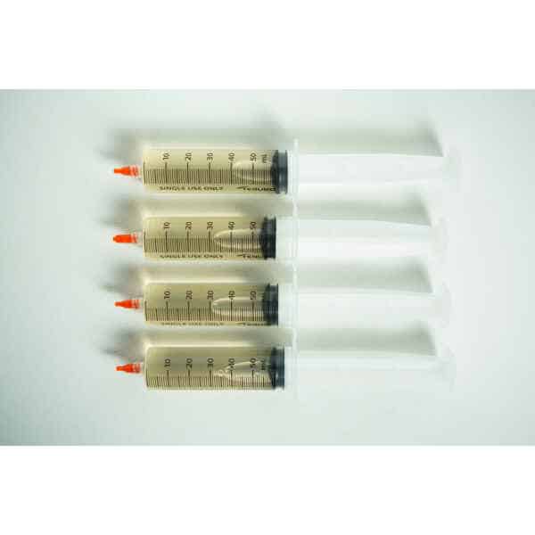 Four large syringes with orange caps from the Liquid culture to Liquid culture expansion Kit are lined up horizontally on a white surface.
