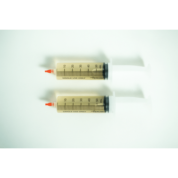 Two large syringes of Liquid culture to Liquid culture expansion Kit, filled with yellowish liquid, are placed side by side on a white surface.