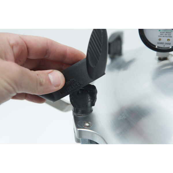 A hand removes a black safety valve from the lid of an All American Tool stainless steel pressure cooker.