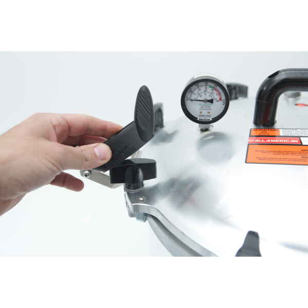 A hand turns the locking mechanism on the lid of an All American Tool pressure cooker with a pressure gauge.