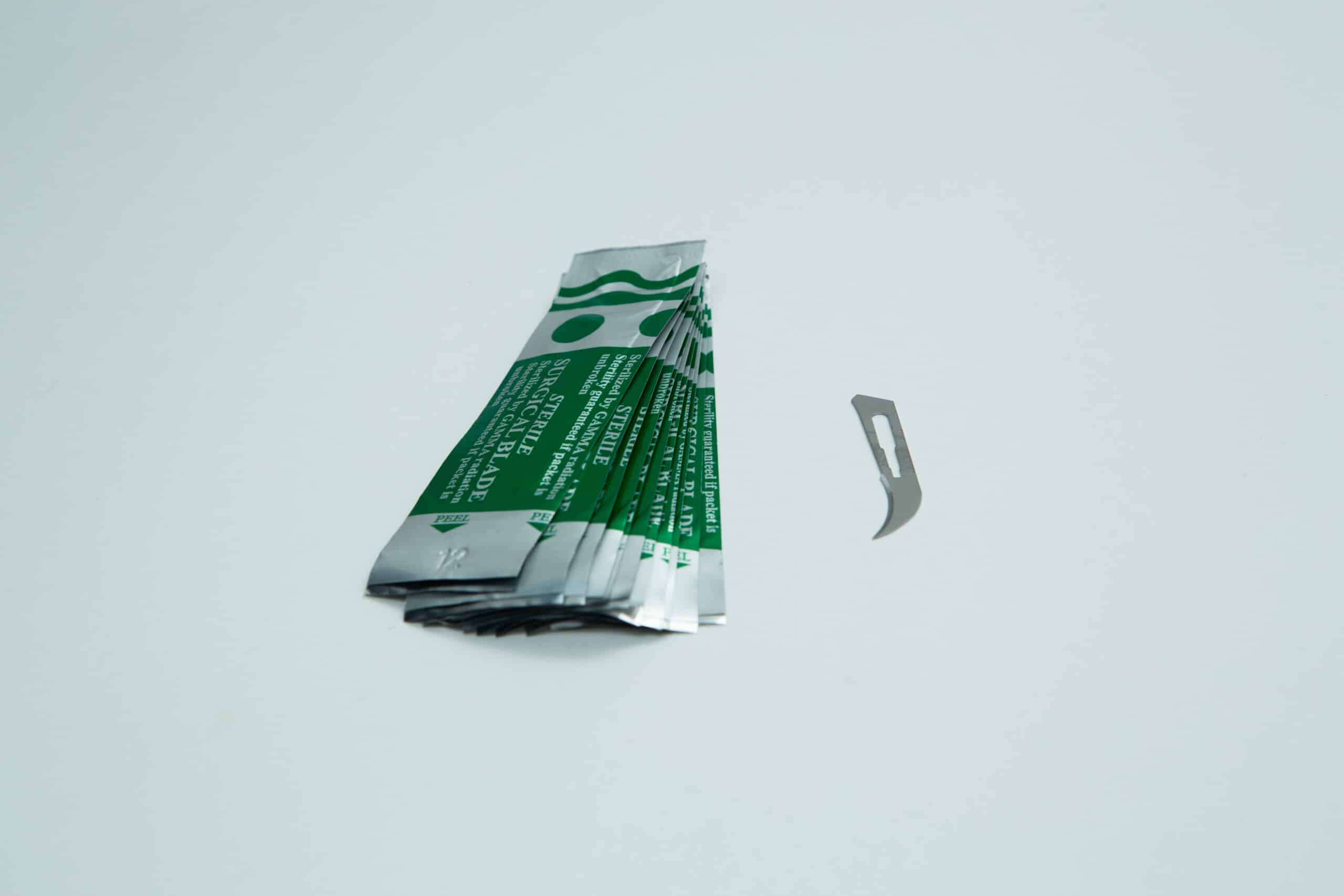 Scalpel handle #3 and blades | Mycology 5 Five green and silver foil packets lie next to a single small, curved metal blade on a white surface.