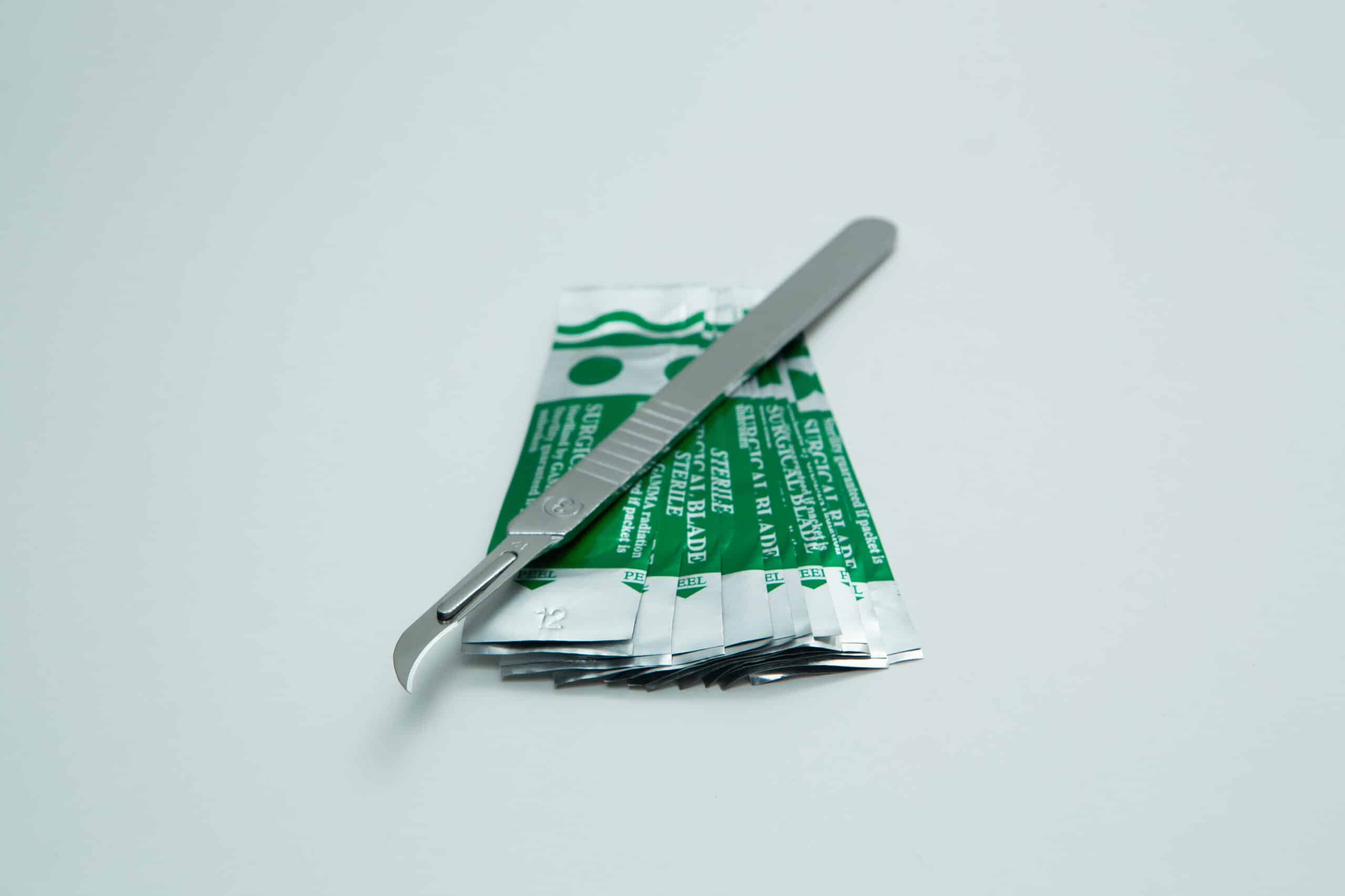 Scalpel handle #3 and blades | Mycology 7 A scalpel rests on several green and silver sterile blade packets on a white background.