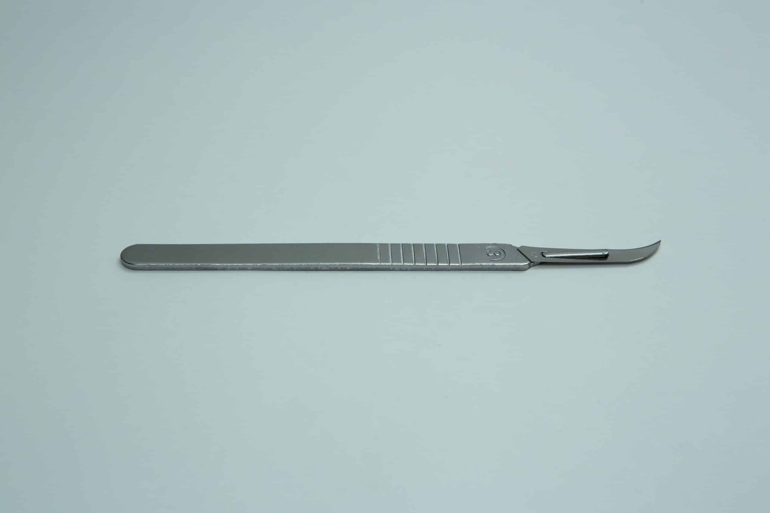 Scalpel handle #3 and blades | Mycology 8 A metal surgical scalpel with a curved blade lies on a plain light grey background.